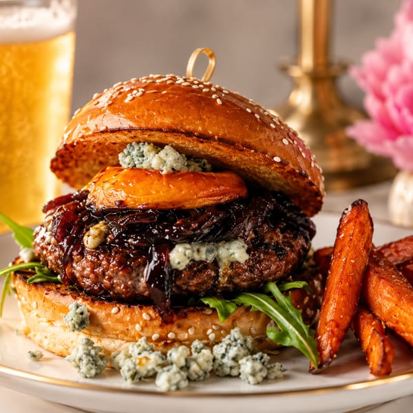Gourmet sesame-seed burger with blue cheese, caramelized onions, and sweet potato fries on a plate beside a beer.