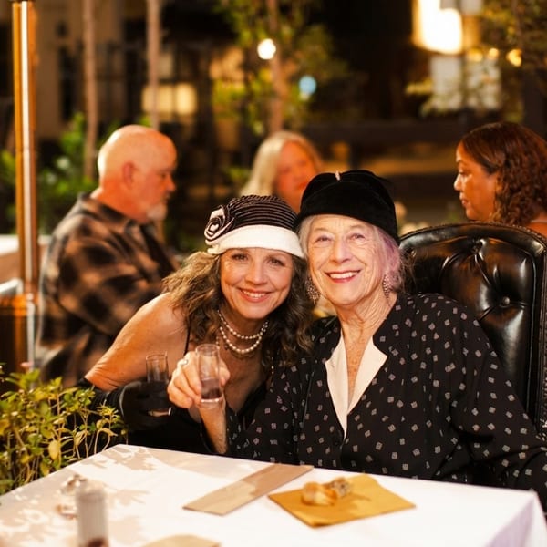 Two smiling women pose at a warmly lit restaurant table, surrounded by diners, drinks, plants, and plated food.
