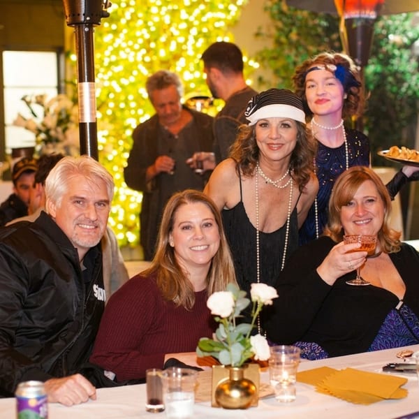 Guests enjoy a festive restaurant gathering, with costumed servers, drinks, appetizers, candlelit tables, and sparkling holiday lights.