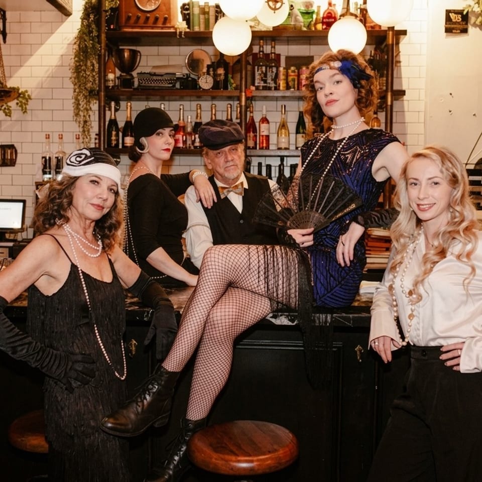 Five people in 1920s-inspired attire pose at a warmly lit restaurant bar surrounded by bottles, shelves, and vintage decor.
