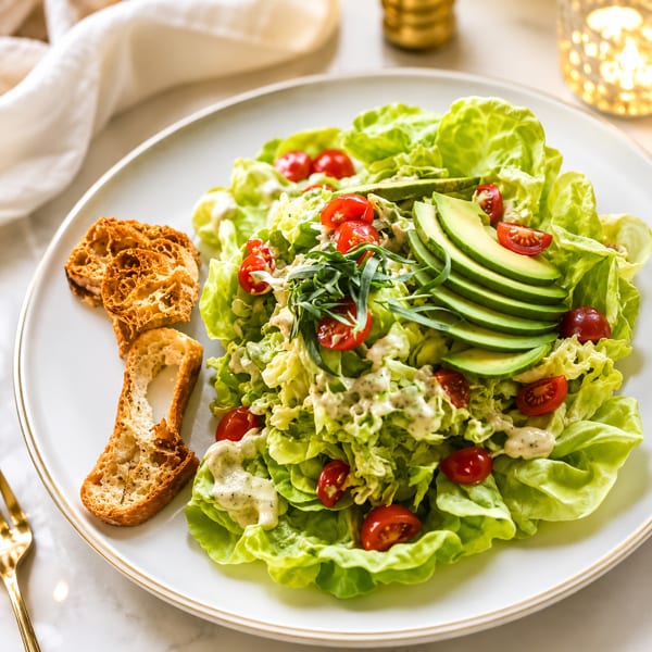 Fresh green salad with avocado, cherry tomatoes, creamy dressing, and toasted bread on a white plate.